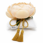 Decoración textil Alba Peonía con flor en tono crema, cojín blanco y borlas doradas sobre fondo claro.