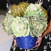 Arreglo floral artificial de gran volumen en tonos verde y crema, sostenido con ambas manos para mostrar su tamaño y su base cilíndrica azul