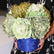 Arreglo floral artificial de gran volumen en tonos verde y crema, sostenido con ambas manos para mostrar su tamaño y su base cilíndrica azul