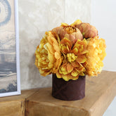 Arreglo floral con peonías y dalias artificiales en tonos amarillos y naranjas sobre una base marrón