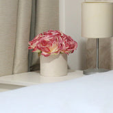 Caja redonda blanca con rosas artificiales rosas y crema, colocada sobre una mesita junto a una lámpara y cortinas beige