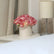 Caja redonda blanca con rosas artificiales rosas y crema, colocada sobre una mesita junto a una lámpara y cortinas beige