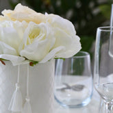 Composición de rosas blancas artificiales en una base cilíndrica blanca con borlas, colocada sobre una mesa preparada para una celebración
