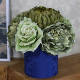 Centro floral artificial con flores verdes y crema en una base cilíndrica azul estampada, colocado sobre un mueble de madera junto a un cuadro