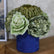 Centro floral artificial con flores verdes y crema en una base cilíndrica azul estampada, colocado sobre un mueble de madera junto a un cuadro
