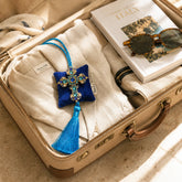Cruz de Amalfi, colgante decorativo artesanal de SimLi Atelier sobre una maleta de viaje con ropa de lino y accesorios de verano.
