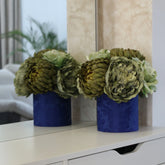Decoración floral artificial en tonos verdes y crema sobre una base azul, colocada delante de un espejo que refleja la parte posterior de la composición