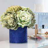 Composición de flores artificiales en tonos verde y crema sobre una base cilíndrica azul, colocada en un tocador junto a productos de maquillaje