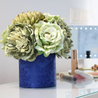 Composición de flores artificiales en tonos verde y crema sobre una base cilíndrica azul, colocada en un tocador junto a productos de maquillaje