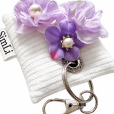 Llavero textil Lila con detalle de flores en tonos lila, perlas y aro metálico sobre fondo blanco.