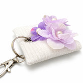 Llavero textil blanco con flores lilas, detalles de perlas y aro metálico sobre fondo blanco.