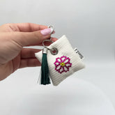 Detalle de un llavero blanco sostenido en la mano con flor de cristales rosas y amarillos y borla verde oscura