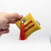 Llavero decorativo amarillo con diseño de la bandera de España en cristales y borla roja, sostenido en la mano.