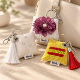 Tres llaveros textiles hechos a mano en forma de cojín: uno blanco con borla blanca y cristales, uno blanco con flor burdeos y perla central, y uno amarillo con cristales rojos y amarillas y borla roja, junto a un bolso beige.