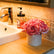 Caja redonda con rosas artificiales rosas y crema colocada sobre una encimera de baño junto a un lavabo y un dispensador de jabón