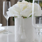 Centro floral con rosas blancas artificiales en una base blanca decorada con borlas, colocado entre copas sobre una mesa