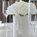 Centro floral con rosas blancas artificiales en una base blanca decorada con borlas, colocado entre copas sobre una mesa