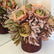 Rosas y crisantemos artificiales en tonos rosa, verde y malva sobre una base decorativa burdeos
